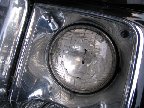 1991 Ford LTD Crown Victoria - damaged round sealed-beam headlamp with a broken lens, set in a chrome bezel on a dark vehicle.
