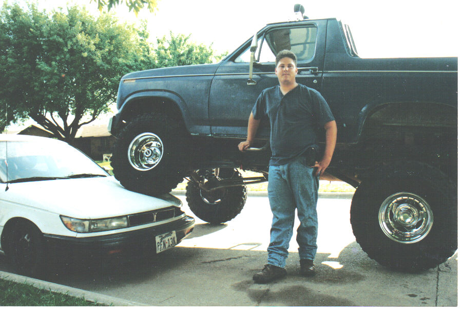 Dark blue lifted Ford Bronco on big tires with man standing beside it in a driveway.
