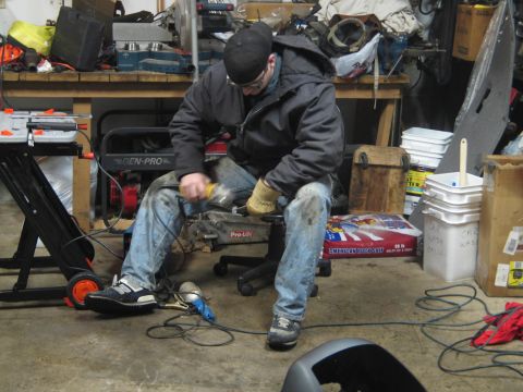 1995 Ford Bronco - A man works on a car part in a cluttered garage with tools and equipment around him.