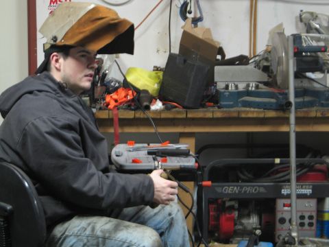 1995 Ford Bronco - A man wearing a welding mask and holding a welding tool sits in a chair in a workshop, with a generator and various tools in the background.