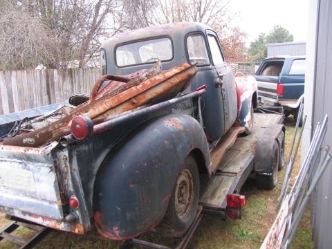 1949 Chevrolet C/K Pickup - A weathered pickup truck, carrying rusty parts, is parked on a trailer next to a fence.