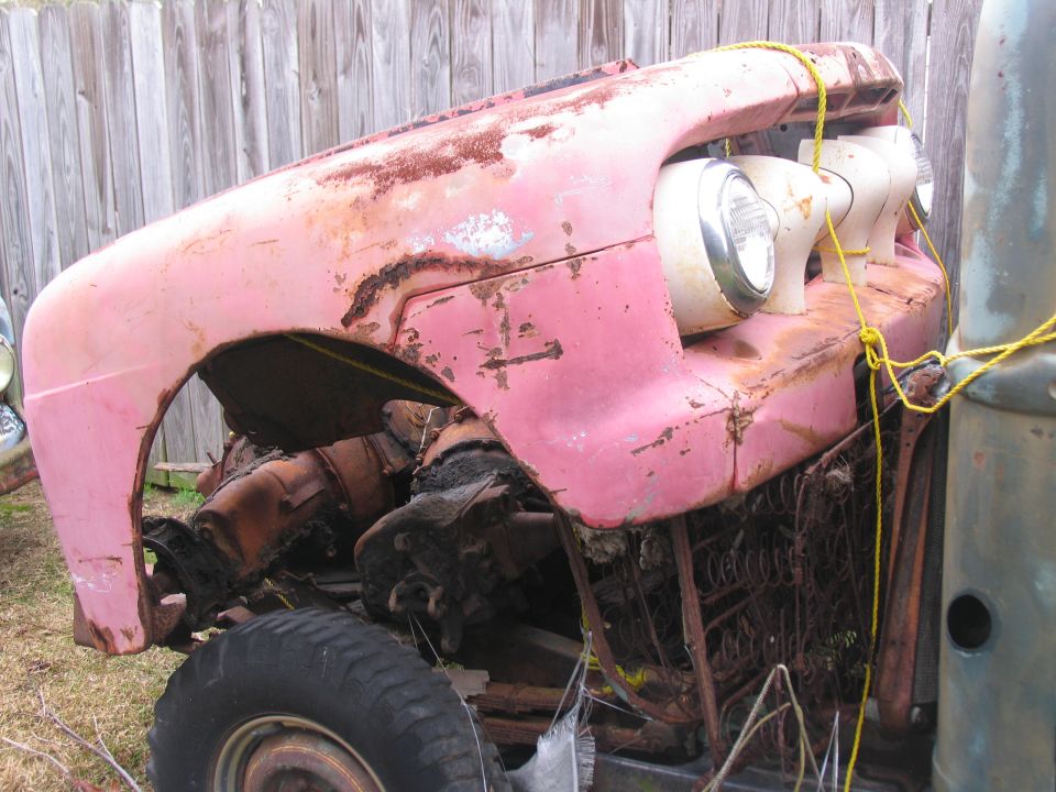 Rusty pink vintage pickup front end, dismantled, tied with yellow rope by a wooden fence