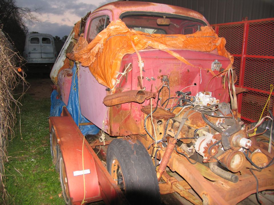 Faded-pink vintage Ford pickup cab on a trailer, stripped front end with exposed rusty engine and wiring.