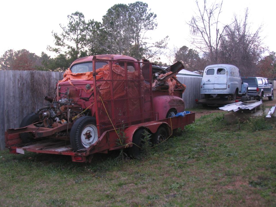 Faded red COE truck being parted out on a red trailer, blue panel van and dark truck behind by a wood fence.