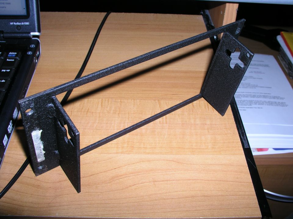 Black metal bracket with mounting holes on a wooden surface next to a laptop