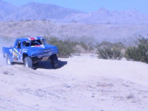 Blue desert race truck driving over sandy terrain with dust trail