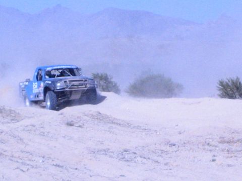 Blue off-road race truck climbing a sandy desert rise in dust