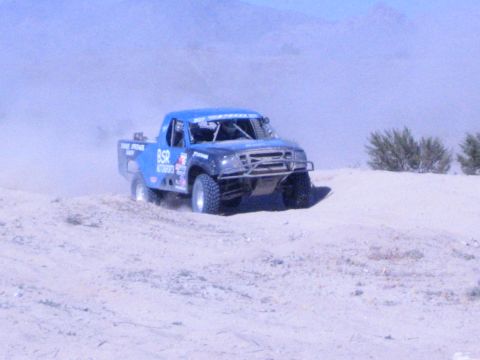 Blue off-road race truck cresting sand dune kicking up dust