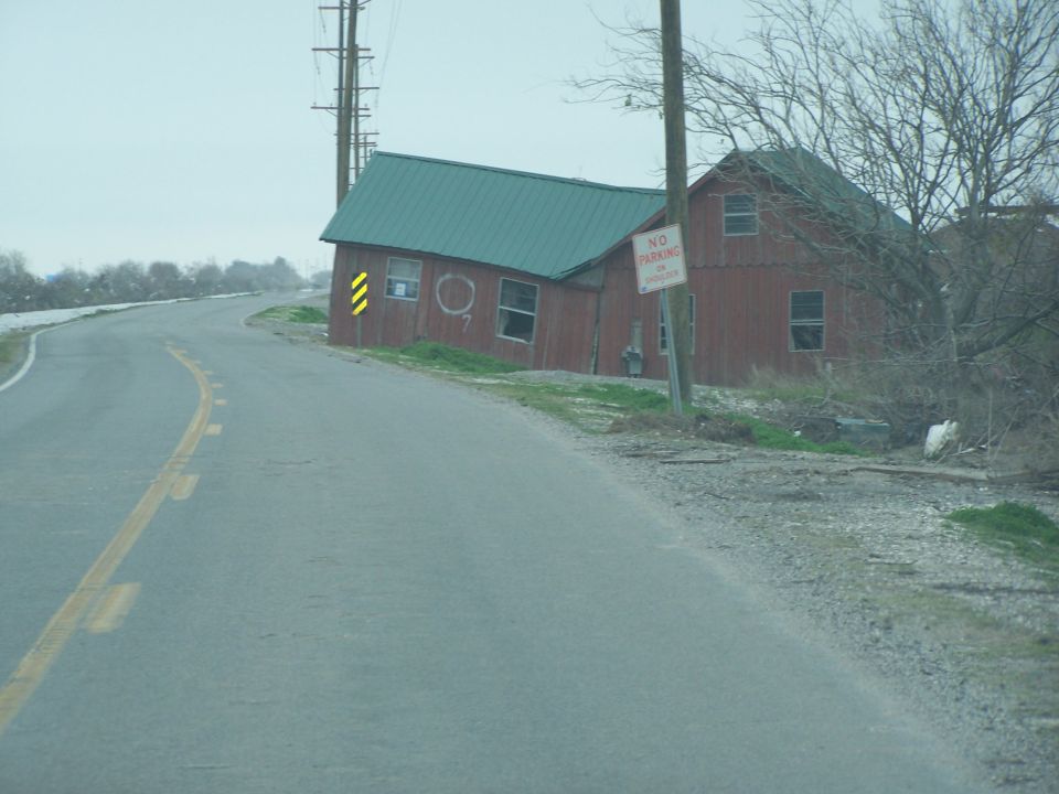 Leaning red barn with green roof next to a road, with a 'No Parking' sign