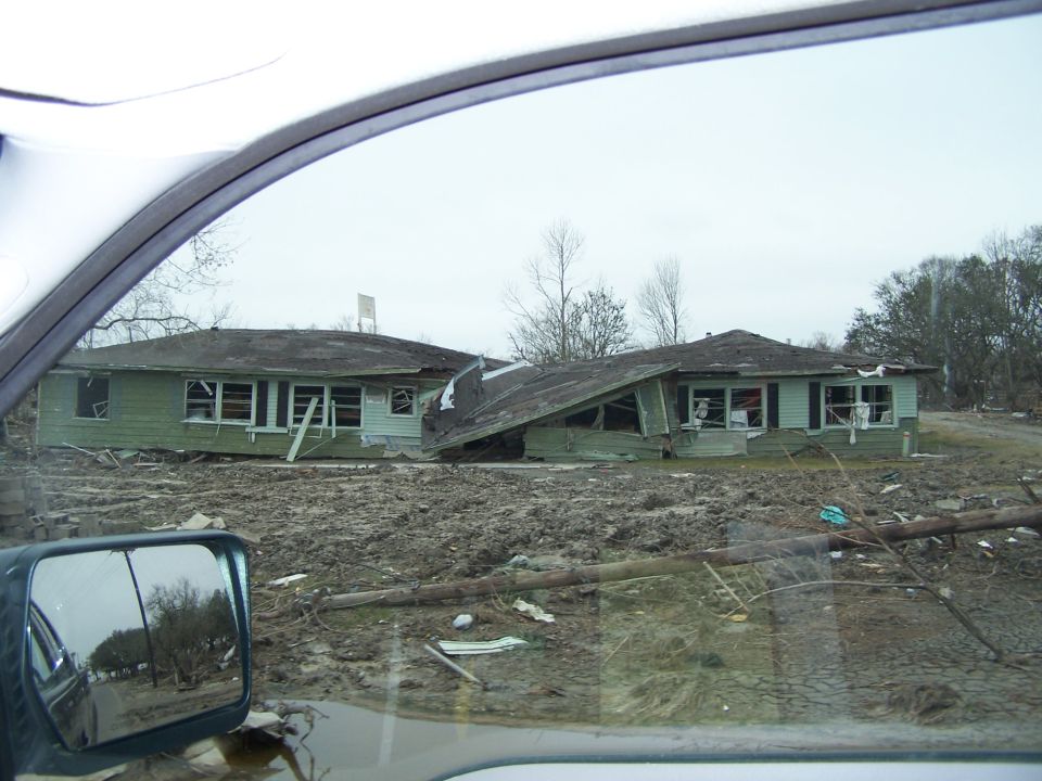 Two houses with damaged roofs and debris scattered around them. View from inside a vehicle