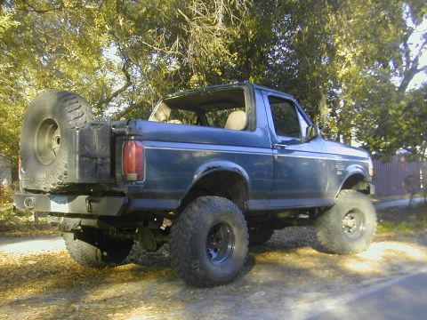 1993 Ford Bronco - Driver-side 3/4 rear view of a lifted blue Ford Bronco with the hardtop removed, parked on a leaf-strewn dirt drive under trees. It carries…