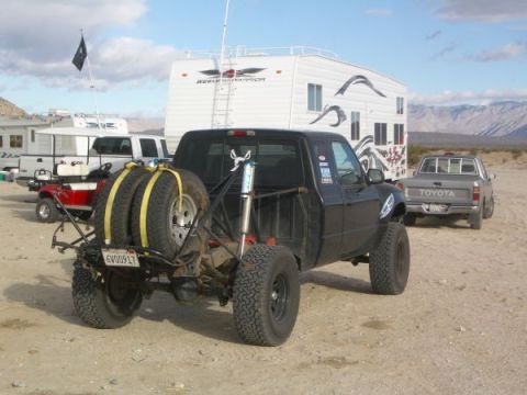 1985 Ford Bronco II - A modified pickup truck is parked in a desert landscape with a trailer and other vehicles nearby.