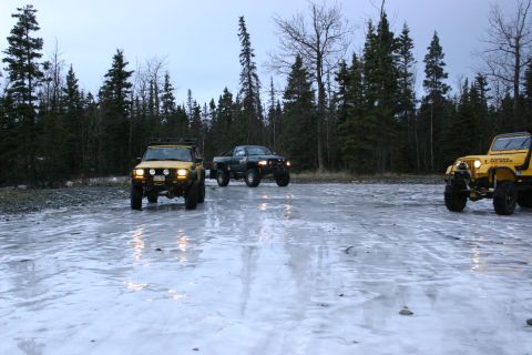 2003 Jeep TJ - Three off-road vehicles are parked on a frozen, reflective surface with a forest in the background.