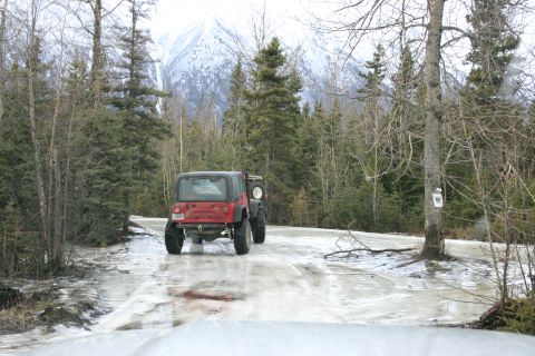 2003 Jeep TJ - A red Jeep Wrangler drives on a partially frozen road surrounded by trees, with a snowy mountain range visible in the distance.