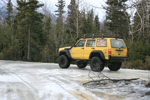 2003 Jeep TJ - Yellow Jeep Cherokee XJ with roof rack and snorkel on an icy road, surrounded by trees.