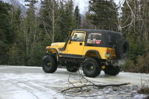 2003 Jeep TJ - A yellow 2003 Jeep TJ with a snorkel and off-road tires navigates an icy surface in a wooded area.