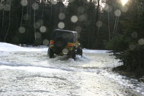 2003 Jeep TJ - A yellow Jeep Wrangler navigates through a water crossing in a wooded area, with snow visible on the banks.