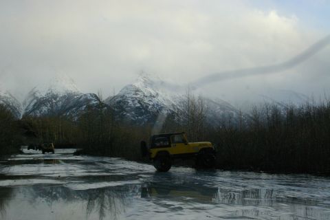 2003 Jeep TJ - Two Jeeps drive on a partially frozen road with snow-capped mountains in the background.