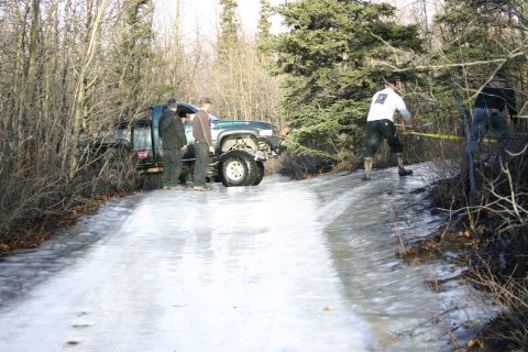 2003 Jeep TJ - A green pickup truck is stuck on an icy road in a wooded area with several people around it.