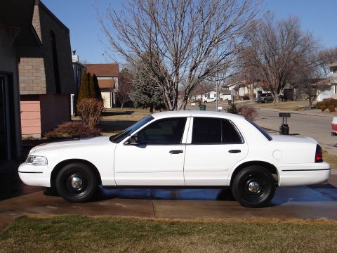 2000 Ford Crown Victoria - A white full-size sedan, consistent with a Ford Crown Victoria, shown in full side profile parked on a concrete driveway. It has black stee…