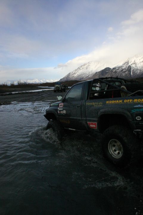 2003 Jeep TJ - Off-road vehicles navigate a riverbed with snow-capped mountains under a cloudy sky.