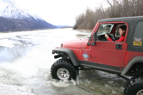 2003 Jeep TJ - A woman drives a red Jeep Wrangler through shallow ice and water near a snow-covered mountain.