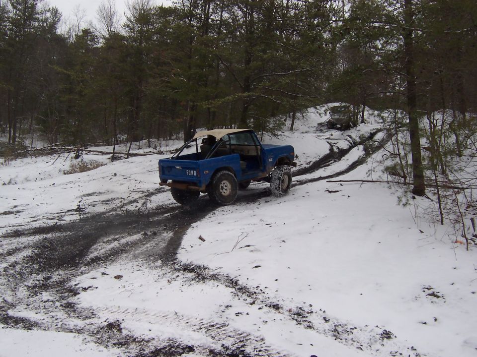 Blue early Ford Bronco descending a snowy wooded trail, rear three-quarter view