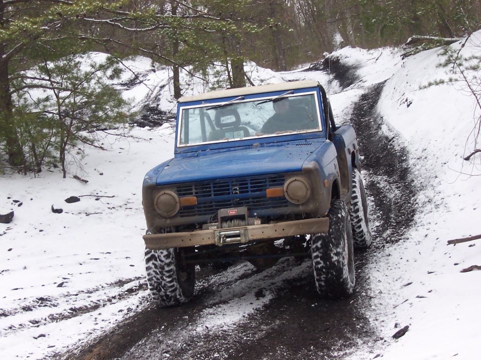 Blue early Ford Bronco with a winch climbing a snowy rutted trail, front view, two occupants