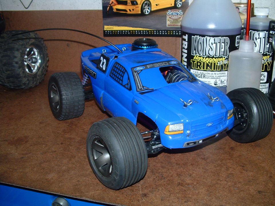 Blue remote control monster truck with large tires, sitting on a brown surface. Bottles of "Monster Horsepower Trinity" fuel are in the background