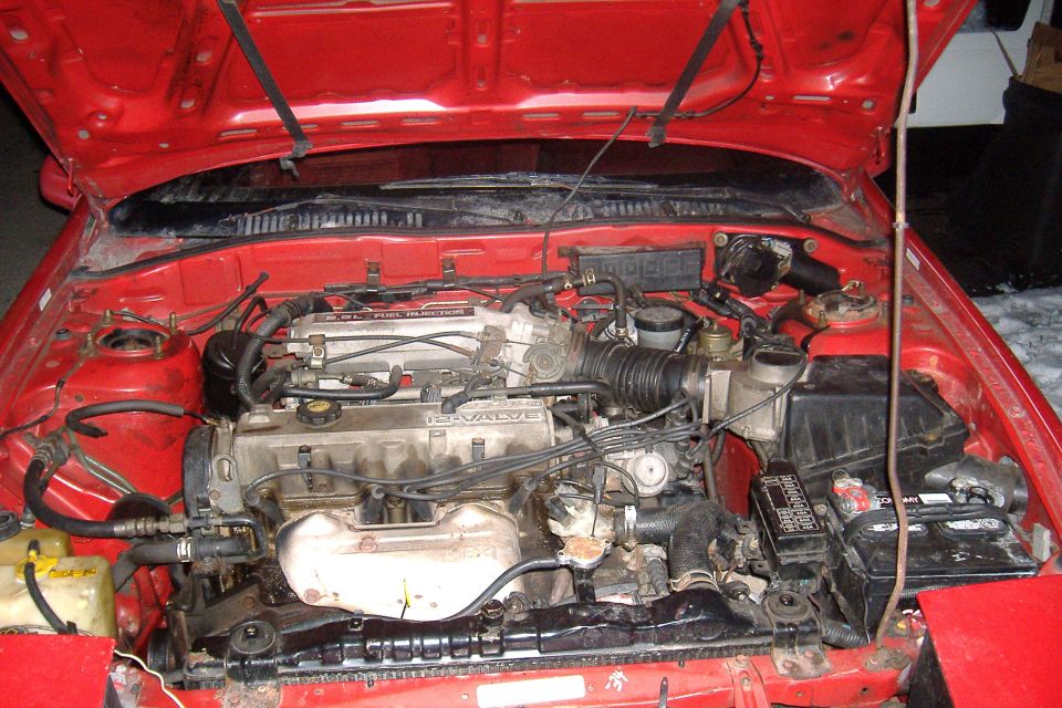 Engine bay of a red car showing a 2.2L 12-valve fuel-injected engine, battery, and intake plumbing