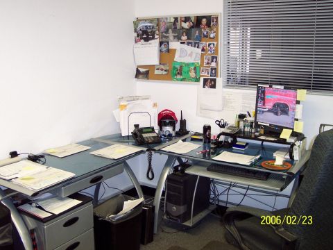 1985 Ford Bronco - A cluttered office desk features a computer screen showing a vehicle, a corkboard with personal photos, and a telephone.