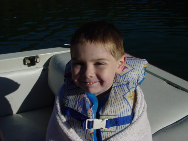 Kid wearing a life jacket and wrapped in a towel sits on a boat. Water is visible in the background
