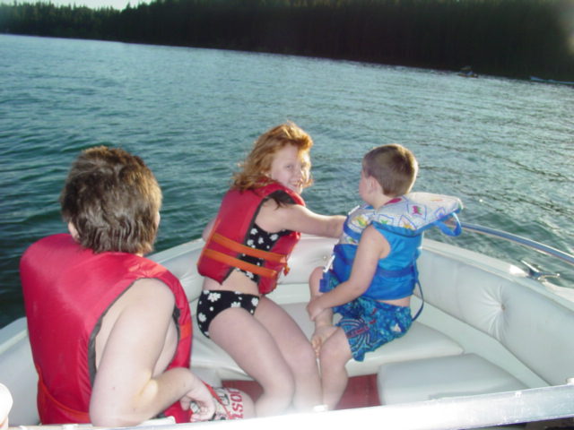 Three children wearing life vests sit in a white boat on a lake