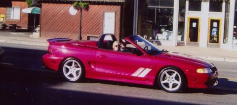 1996 Ford Probe - A pink convertible sports car is seen driving down a street with buildings in the background.