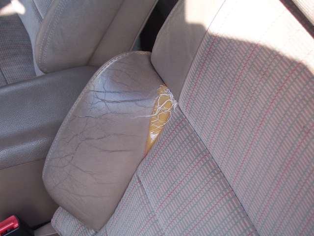 Driver's seat with cracked leather and a large tear showing yellow padding. Fabric seatback has a red and blue striped pattern
