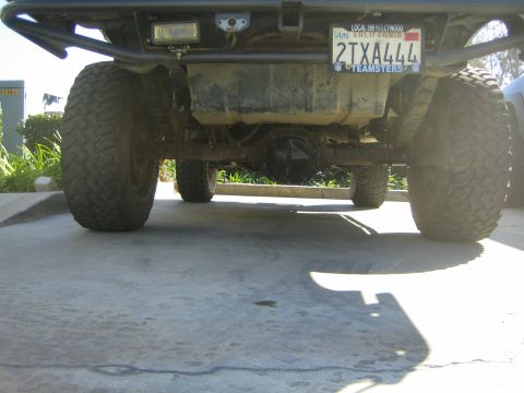 1990 Ford Bronco - Low rear view beneath a lifted truck showing the rear axle, oversized tires, and a California license plate reading 2TXA444.