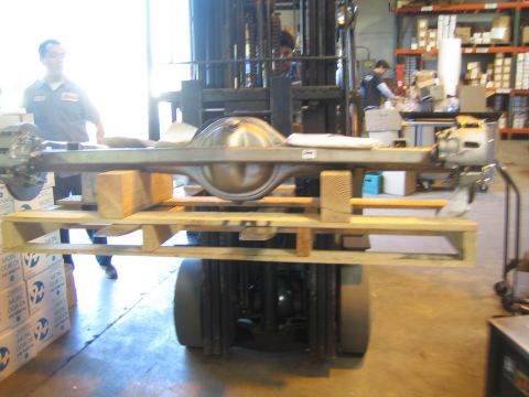 1990 Ford Bronco - forklift carries a large bare axle housing on a pallet while workers move about a busy parts warehouse.