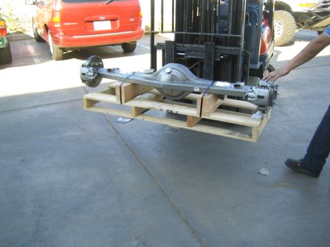 1990 Ford Bronco - bare axle assembly on a wooden pallet is lifted by a forklift in a paved lot, with a red minivan parked nearby.