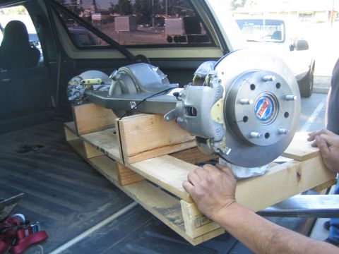 1990 Ford Bronco - person steadies an axle assembly with disc brake rotor on a wooden pallet loaded in the rear of a vehicle.