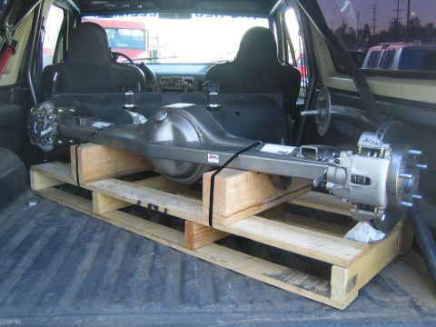 1990 Ford Bronco - fully assembled solid rear axle with differential and disc-brake hub sits strapped to a wooden pallet in a pickup bed.