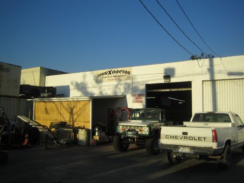 1990 Ford Bronco - Exterior of the Four X Doctor 4WD repair shop at dusk with an early Ford Bronco and a white Chevrolet pickup parked out front.