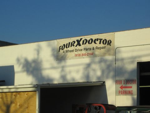 1990 Ford Bronco - vinyl banner reading 'Four X Doctor 4 Wheel Drive Parts & Repair' with a parking arrow on a sunlit white building.
