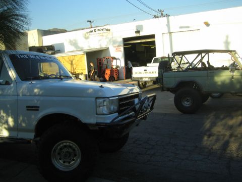 1990 Ford Bronco - lifted white Ford pickup and a white early Bronco parked outside the Four X Doctor shop with a Chevrolet truck between them.