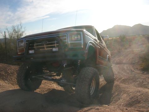 1984 Ford Bronco - This lifted Ford Bronco is navigating a dusty, off-road trail under a bright sky.
