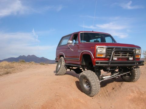 1984 Ford Bronco - A red Ford Bronco is parked on a dirt path with mountains in the background under a blue sky.