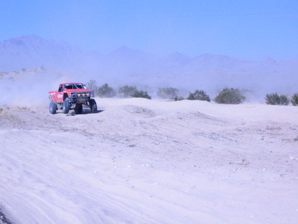 Red off-road race truck cresting a sandy berm with mountains in the background