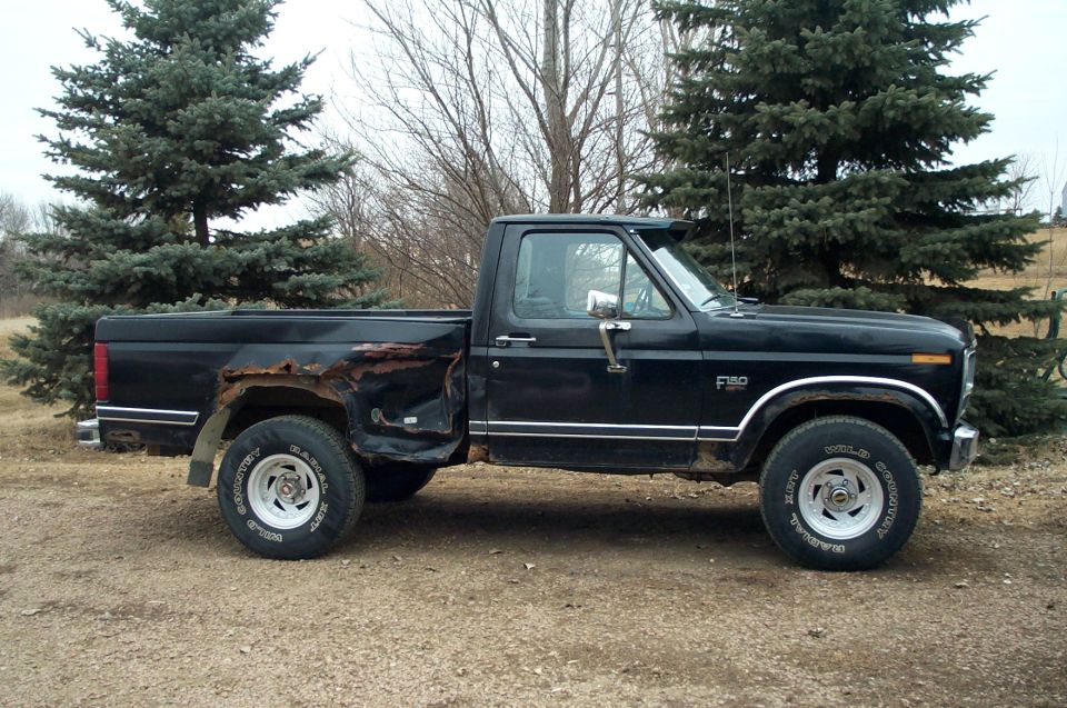 Black Ford F-150 pickup side view with rust damage on rear fender, parked on gravel near pine trees