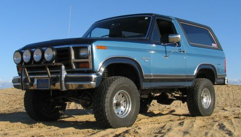 1984 Ford Bronco - This lifted Ford Bronco sits on a sandy terrain under a clear blue sky.