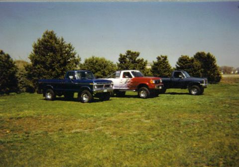 1986 Ford Bronco - Three pickup trucks are parked side-by-side on a grassy lawn with trees in the background.