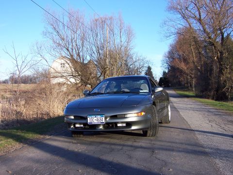 1996 Ford Probe - A gray Ford Probe is parked on the side of a road with trees and a house in the background.